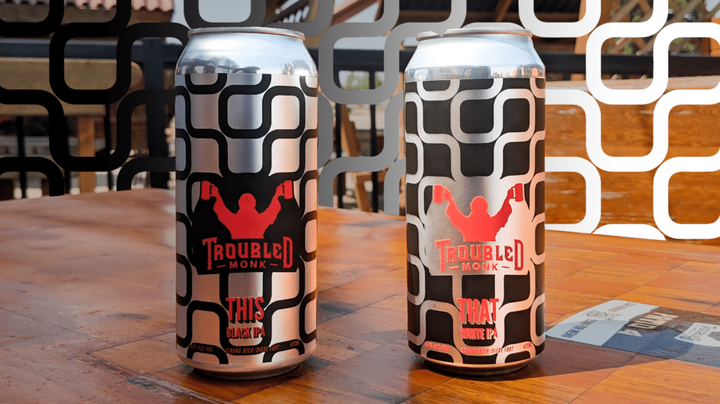 A can of THIS White IPA in silver and black, and a cna of THAT Black IPA in black and silver sitting on a table at troubled monk taproom 