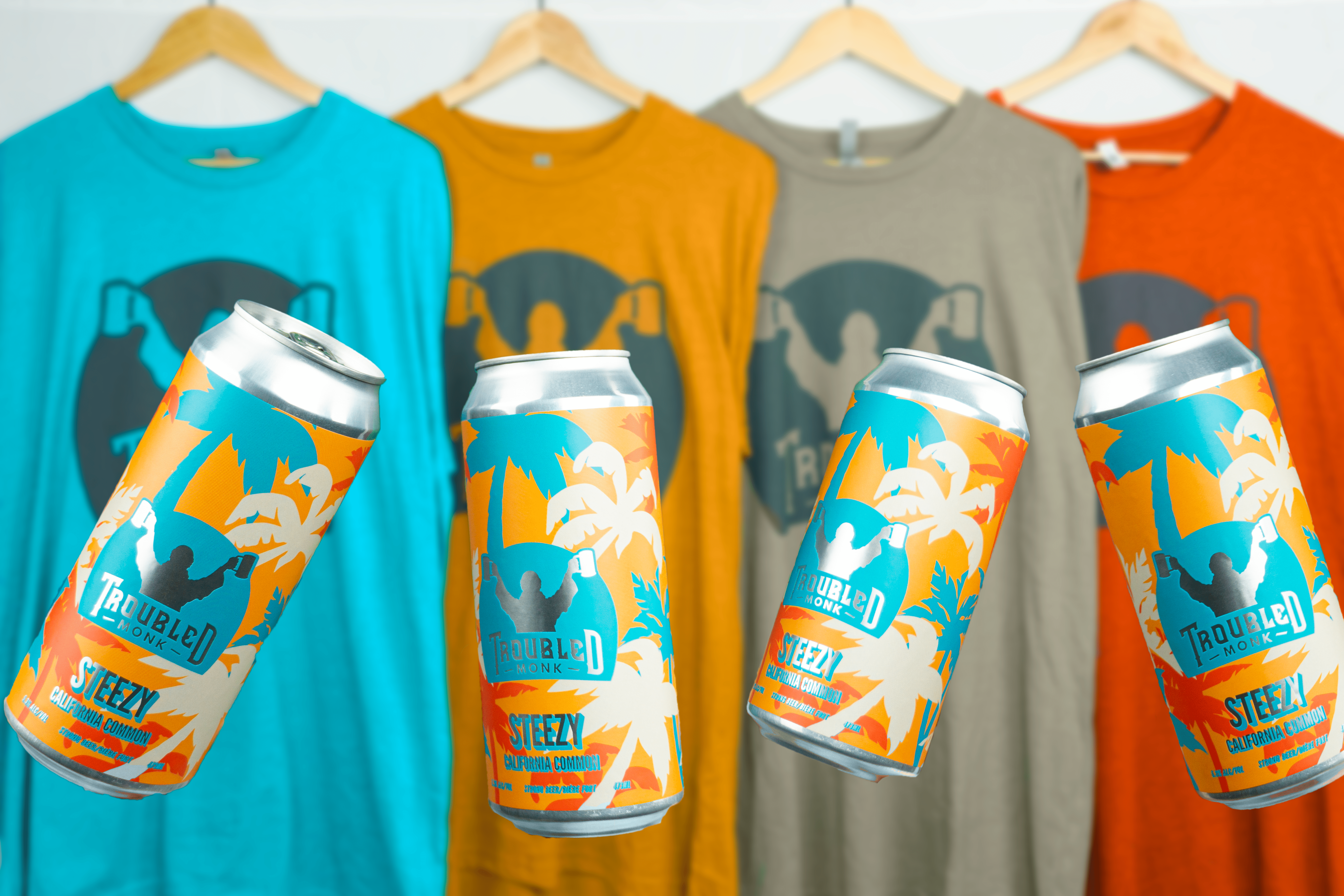 Steezy cans next to Troubled Monk shirts in turquoise, pumpkin orange, brown, and bright orange
