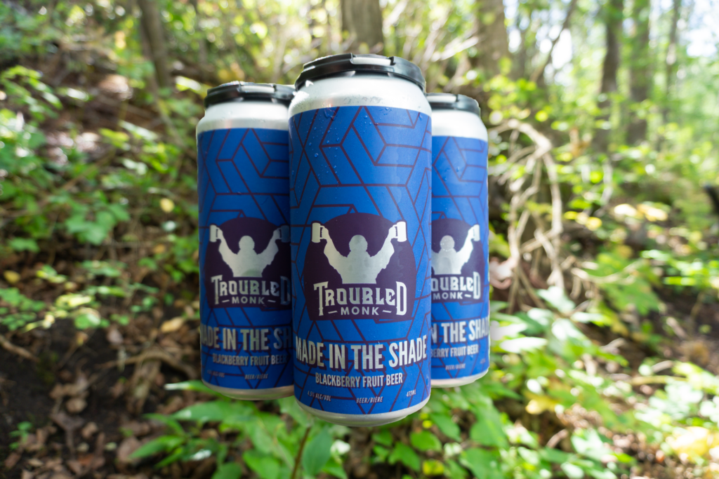 four black and blue tallboy cans made in the shade blackberry fruit beer under the shade of the trees behind Troubled Monk