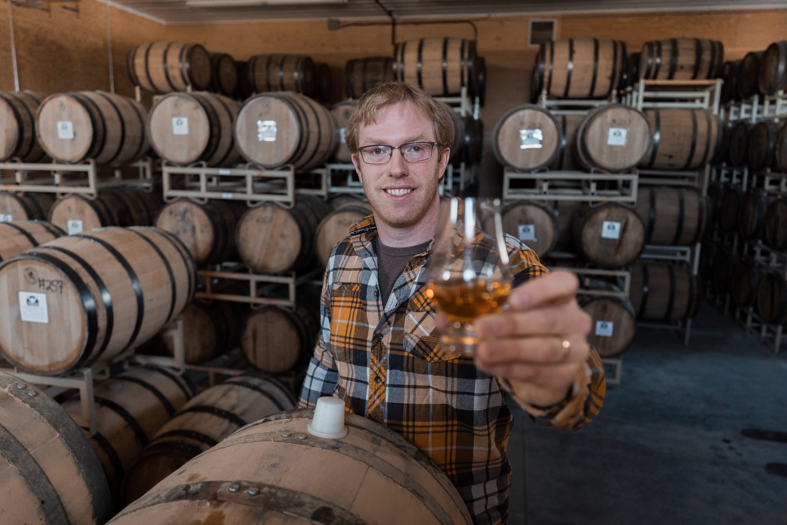 picture of garret haynes head distiller holding a glass of meeting creek single malt whisky up with the barrels of whisky all around him